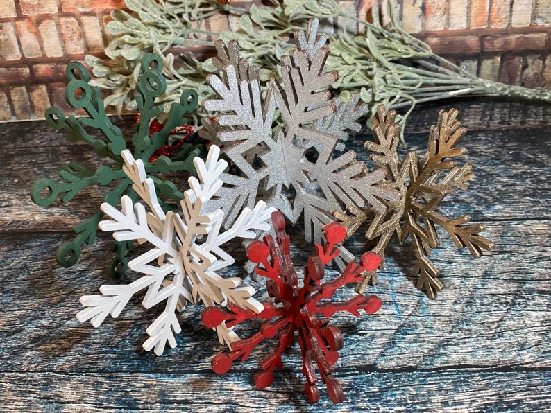 May include: Six wooden snowflake ornaments in various colors, including green, silver, white, gold, and red. The snowflakes are arranged on a rustic wooden surface.