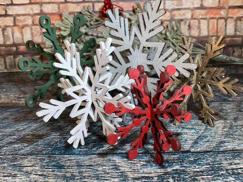 May include: Five wooden snowflake ornaments in various colors, including green, silver, white, gold, and red. The snowflakes are layered on top of each other and are arranged on a wooden surface.