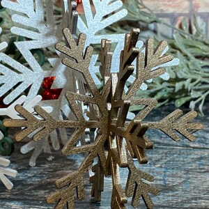 May include: A gold glitter snowflake ornament with a 3D design. The snowflake is made of wood and has a rustic look.
