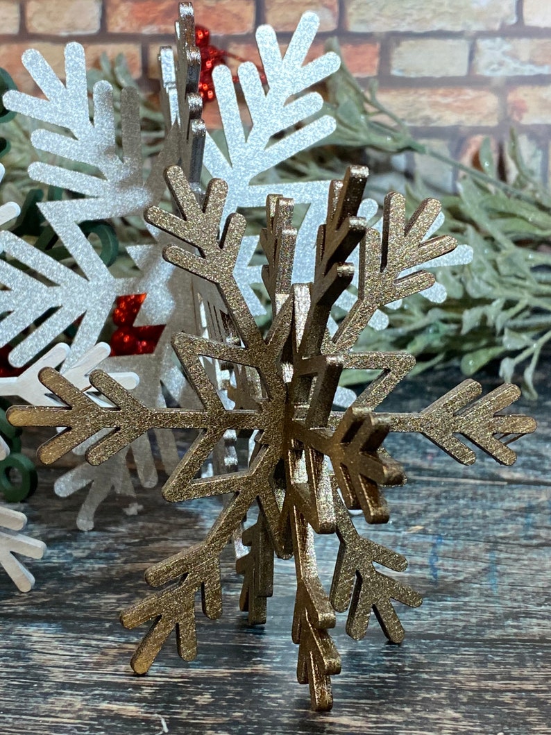May include: A gold glitter snowflake ornament with a 3D design. The snowflake is made of wood and has a rustic look.