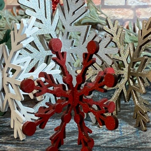 May include: A set of three wooden snowflakes, one red, one silver, and one gold. The snowflakes are all intricately cut and have a rustic, vintage look.