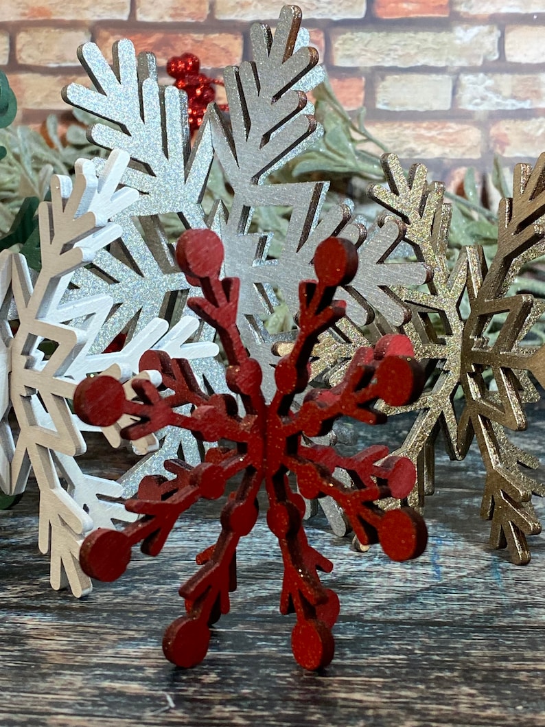 May include: A set of three wooden snowflakes, one red, one silver, and one gold. The snowflakes are all intricately cut and have a rustic, vintage look.