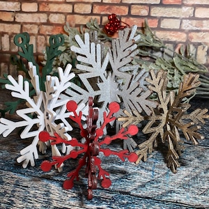 May include: Five wooden snowflake ornaments in various colors, including silver, gold, white, and red. The snowflakes are arranged in a cluster on a wooden surface.