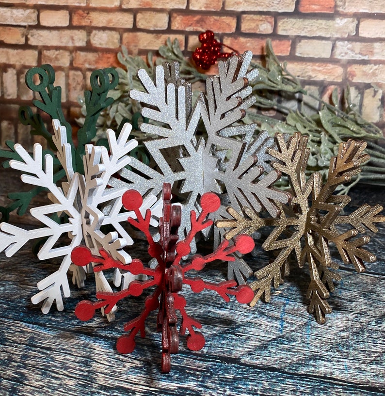 May include: Five wooden snowflake ornaments in various colors, including silver, gold, white, and red. The snowflakes are arranged in a cluster on a wooden surface.