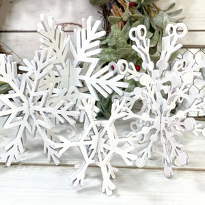 May include: A set of five white wooden snowflakes of varying sizes. The snowflakes are layered on top of each other, creating a three-dimensional effect.