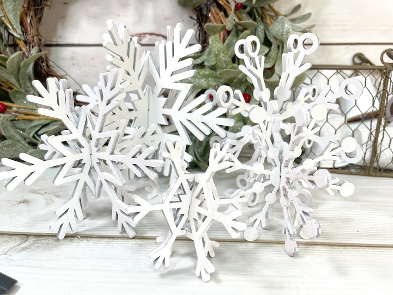 May include: A set of five white wooden snowflakes of varying sizes. The snowflakes are layered on top of each other, creating a three-dimensional effect.