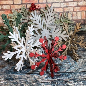 May include: Five decorative wooden snowflakes in various colors, including green, silver, gold, white, and red. The snowflakes are arranged in a cluster on a wooden surface.