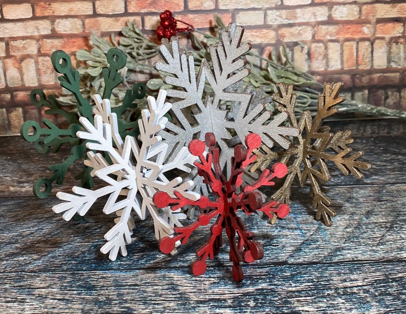 May include: Five decorative wooden snowflakes in various colors, including green, silver, gold, white, and red. The snowflakes are arranged in a cluster on a wooden surface.