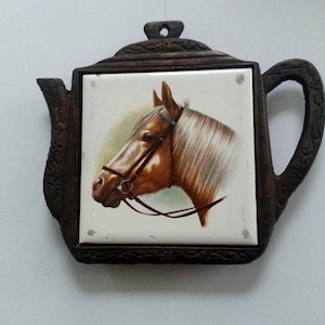 Puede incluir: Un calienta-tetera de metal marrón y negro con una baldosa de cerámica blanca que presenta un caballo marrón con una brida azul. El caballo está mirando hacia la izquierda y tiene una crin y una cola blancas.