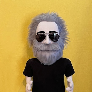 May include: A gray-haired puppet wearing black sunglasses and a black t-shirt. The puppet has a gray beard and mustache. The puppet is standing on a wooden base.