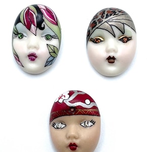 May include: Three oval-shaped ceramic face cabochons. The top left features green eyes and pink floral hair accents. The top right has orange eyes and a leopard print headpiece. The bottom face has clear crystal eyes and a red patterned headpiece.