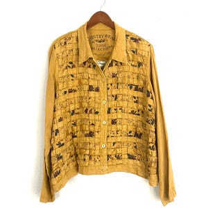 May include: Mustard yellow button-down jacket with long sleeves. The front features a grid pattern with floral accents. The collar has the text "COUNTRY WEAR 2000 COLLECTIONS". The jacket is hanging on a wooden hanger.