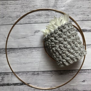 Puede incluir: Un macetero de hormigón gris con forma de calavera con flores blancas en su interior. El macetero está sobre una superficie de madera blanca con un anillo de metal dorado a su alrededor.
