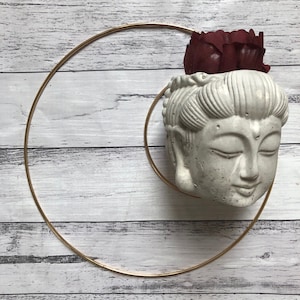 Puede incluir: Un macetero de cabeza de Buda de hormigón blanco con una flor burdeos en la parte superior. El macetero está rodeado por un anillo de metal dorado.