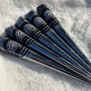 Peut inclure: Six cônes de henné noirs, chacun enveloppé dans du plastique transparent. Chaque cône porte l'inscription "ARTIST'S" et "ORGANIC JAGUA". Les cônes sont disposés sur une surface blanche et duveteuse.