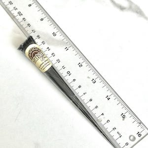 Pode incluir: Um cone de henna preto com o texto "ARTISTIC Henna" e um número de telefone no rótulo. O cone está sobre uma superfície branca ao lado de uma régua.