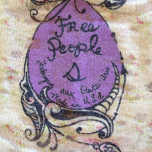 K&ouml;nnte beinhalten: Ein lilafarbenes Etikett mit dem Text "Free People" und einer Gr&ouml;&szlig;e "S" in schwarzer Schrift. Das Etikett ist von einem schwarzen floralen Design auf cremefarbenem Hintergrund umgeben.