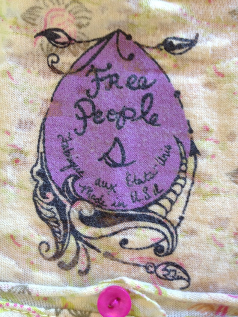 K&ouml;nnte beinhalten: Ein lilafarbenes Etikett mit dem Text "Free People" und einer Gr&ouml;&szlig;e "S" in schwarzer Schrift. Das Etikett ist von einem schwarzen floralen Design auf cremefarbenem Hintergrund umgeben.