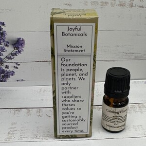 Op de afbeelding: Een kleine bruine glazen fles met een zwarte dop met het label "Calming Focus" naast een doos met de tekst "Joyful Botanicals". De doos heeft een missieverklaring over duurzame inkoop. De achtergrond bevat lavendeltakken.