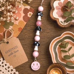 May include: A beaded charm featuring pink and black mushroom beads, pink beads, and a smiley face charm. The charm is strung on a clear cord. A tag that says "HANDMADE with love" is also visible.