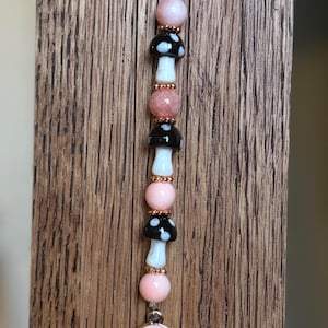 May include: A vertical beaded charm featuring alternating light pink spherical beads and black mushroom-shaped beads with white spots. Small metallic rose gold spacers separate the beads. A round light pink smiley face charm hangs at the bottom.