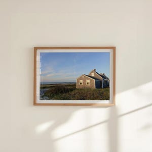 K&ouml;nnte beinhalten: Gerahmter Druck einer K&uuml;stenlandschaft mit einem verwitterten grauen Haus mit mehreren Fenstern. Das Haus steht vor einem blauen Himmel, dem Meer und grasbewachsenen D&uuml;nen. Der Holzrahmen verleiht dem Kunstwerk eine nat&uuml;rliche Note.