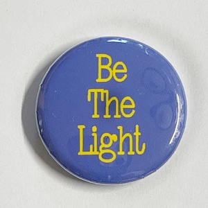 May include: A purple button with the text "Be The Light" in yellow lettering.
