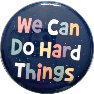 May include: A navy blue button with the text "We can do hard things" in colorful letters. The letters are outlined in white and have a speckled texture.