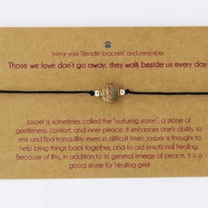 May include: A bracelet featuring a central jasper bead and black cord, presented on a brown card. The card includes text about the jasper stone and a message about remembrance.