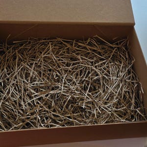 May include: An open, rectangular cardboard box filled with shredded paper. The box is a light brown color, and the paper is a mix of light and medium brown tones. The box is empty and ready for packaging.