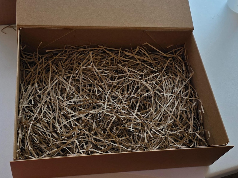May include: An open, rectangular cardboard box filled with shredded paper. The box is a light brown color, and the paper is a mix of light and medium brown tones. The box is empty and ready for packaging.