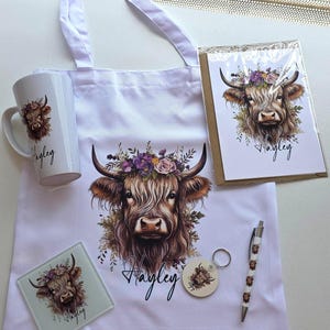 May include: A collection of white items featuring a Highland cow design with a floral crown. Includes a tote bag, mug, greeting card, coaster, keychain, and pen. The name "Hayley" is written on several items.