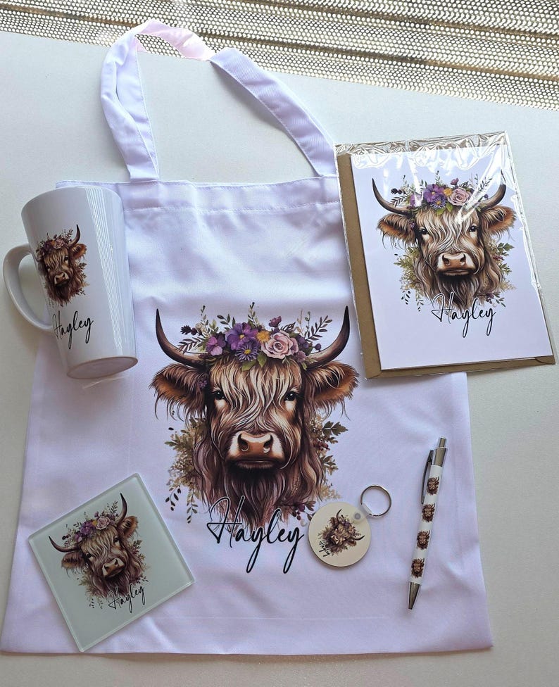 May include: A collection of white items featuring a Highland cow design with a floral crown. Includes a tote bag, mug, greeting card, coaster, keychain, and pen. The name "Hayley" is written on several items.