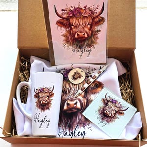 May include: A gift box containing a mug, card, coaster, pen, and tea towel, all featuring a Highland cow illustration with floral accents. The name "Hayley" is written on several items. The items are white with brown, purple, and green details.