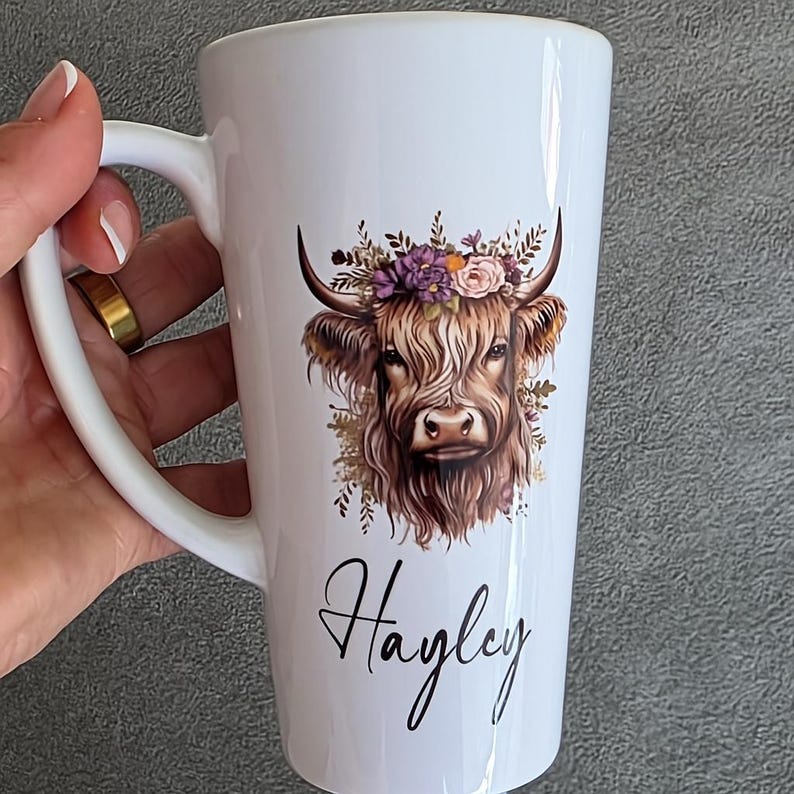 May include: White ceramic mug featuring a Highland cow illustration with a floral crown. The cow has long, shaggy brown fur and is adorned with purple and pink flowers. The name "Hayley" is written in cursive script below the cow.