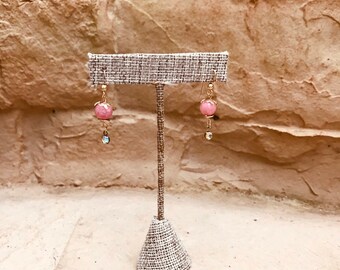 Puede incluir: Un par de pendientes de piedra rosa con detalles dorados colgando de un soporte de tela. Los pendientes tienen una pequeña piedra rosa y un cristal transparente.