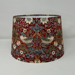 May include: A cylindrical lampshade with a botanical pattern. The design includes birds, flowers, and berries in shades of red, green, cream, and grey. The lampshade has a tapered shape and is suitable for a table or floor lamp.