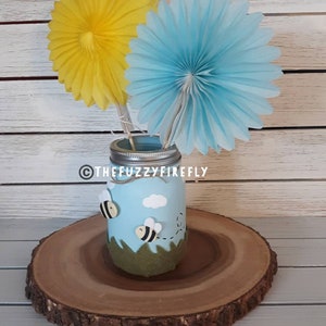 Peut inclure: Un bocal en verre bleu clair avec un couvercle argent&eacute;, orn&eacute; de deux petites abeilles, d'un nuage blanc et d'herbe en feutrine verte. Deux &eacute;ventails en papier jaune et bleu clair sont ins&eacute;r&eacute;s. Le texte THEFUZZYFIREFLY est inscrit sur le bocal.
