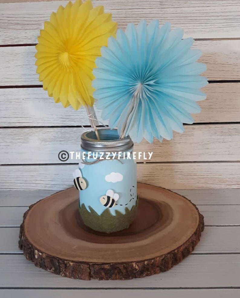 Peut inclure: Un bocal en verre bleu clair avec un couvercle argent&eacute;, orn&eacute; de deux petites abeilles, d'un nuage blanc et d'herbe en feutrine verte. Deux &eacute;ventails en papier jaune et bleu clair sont ins&eacute;r&eacute;s. Le texte THEFUZZYFIREFLY est inscrit sur le bocal.