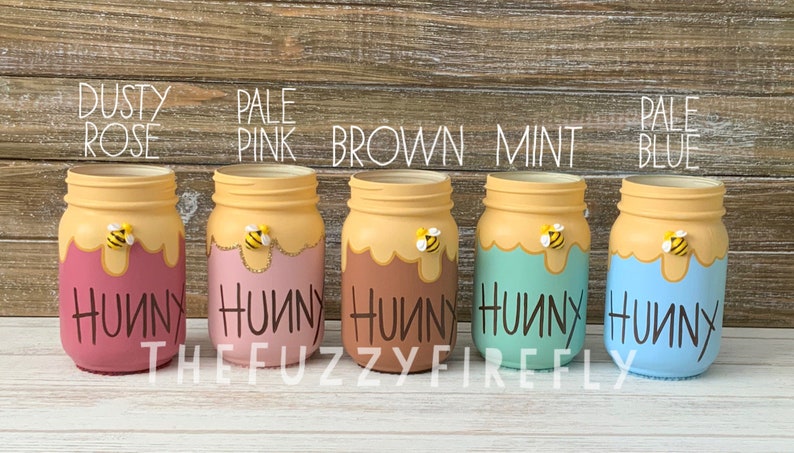 Peut inclure: Cinq bocaux en verre peints en vieux rose, rose p&acirc;le, marron, menthe et bleu p&acirc;le. Chaque bocal pr&eacute;sente un motif de coul&eacute;e de miel jaune sur le dessus, une petite figurine d'abeille et le mot "HUNNY" &eacute;crit en noir sur le devant.
