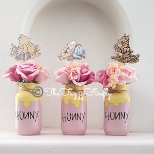 Peut inclure: Trois pots en verre rose avec du miel jaune qui coule sur les c&ocirc;t&eacute;s. Chaque pot porte le mot "Hunny" &eacute;crit en noir et est d&eacute;cor&eacute; d'un personnage de Winnie l'ourson. Les pots sont remplis de fleurs roses et blanches.