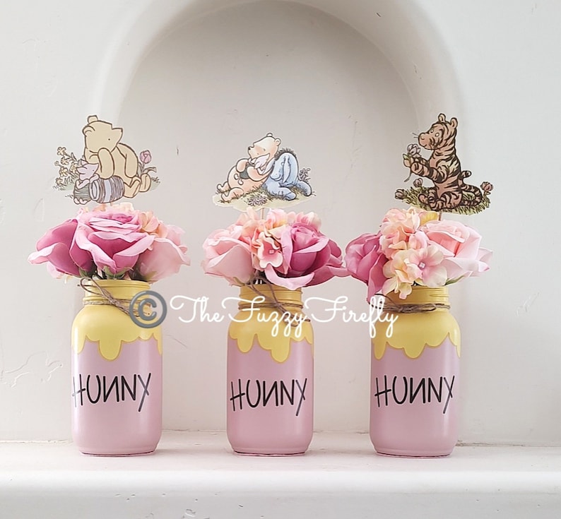 Peut inclure: Trois pots en verre rose avec du miel jaune qui coule sur les c&ocirc;t&eacute;s. Chaque pot porte le mot "Hunny" &eacute;crit en noir et est d&eacute;cor&eacute; d'un personnage de Winnie l'ourson. Les pots sont remplis de fleurs roses et blanches.