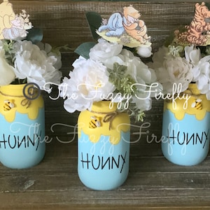 Peut inclure: Trois pots en verre d&eacute;cor&eacute;s sur le th&egrave;me de Winnie l'ourson. Chaque pot est peint en bleu avec du miel jaune qui coule sur le c&ocirc;t&eacute;. Les pots ont une abeille et un personnage de Winnie l'ourson sur le dessus. Les pots sont remplis de fleurs blanches et sont pos&eacute;s sur une surface en bois. Les pots indiquent "Hunny" sur le devant.