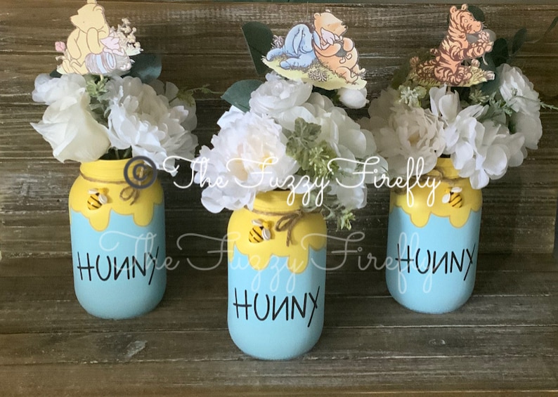 Peut inclure: Trois pots en verre d&eacute;cor&eacute;s sur le th&egrave;me de Winnie l'ourson. Chaque pot est peint en bleu avec du miel jaune qui coule sur le c&ocirc;t&eacute;. Les pots ont une abeille et un personnage de Winnie l'ourson sur le dessus. Les pots sont remplis de fleurs blanches et sont pos&eacute;s sur une surface en bois. Les pots indiquent "Hunny" sur le devant.
