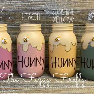 Peut inclure: Quatre pots en verre peints dans diff&eacute;rentes couleurs avec un motif de miel qui coule et une abeille sur le dessus. Les pots sont &eacute;tiquet&eacute;s "Hunny" en lettres noires. Les couleurs sont rose poussi&eacute;reux, p&ecirc;che, jaune soleil et sauge.