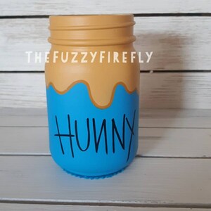 Peut inclure: Un bocal en verre peint avec une section sup&eacute;rieure beige rappelant du miel qui coule et une section inf&eacute;rieure bleu vif. Le mot HUNNY est &eacute;crit en noir dans une police manuscrite sur la partie bleue. Le texte THEFUZZYFIREFLY est visible au-dessus.