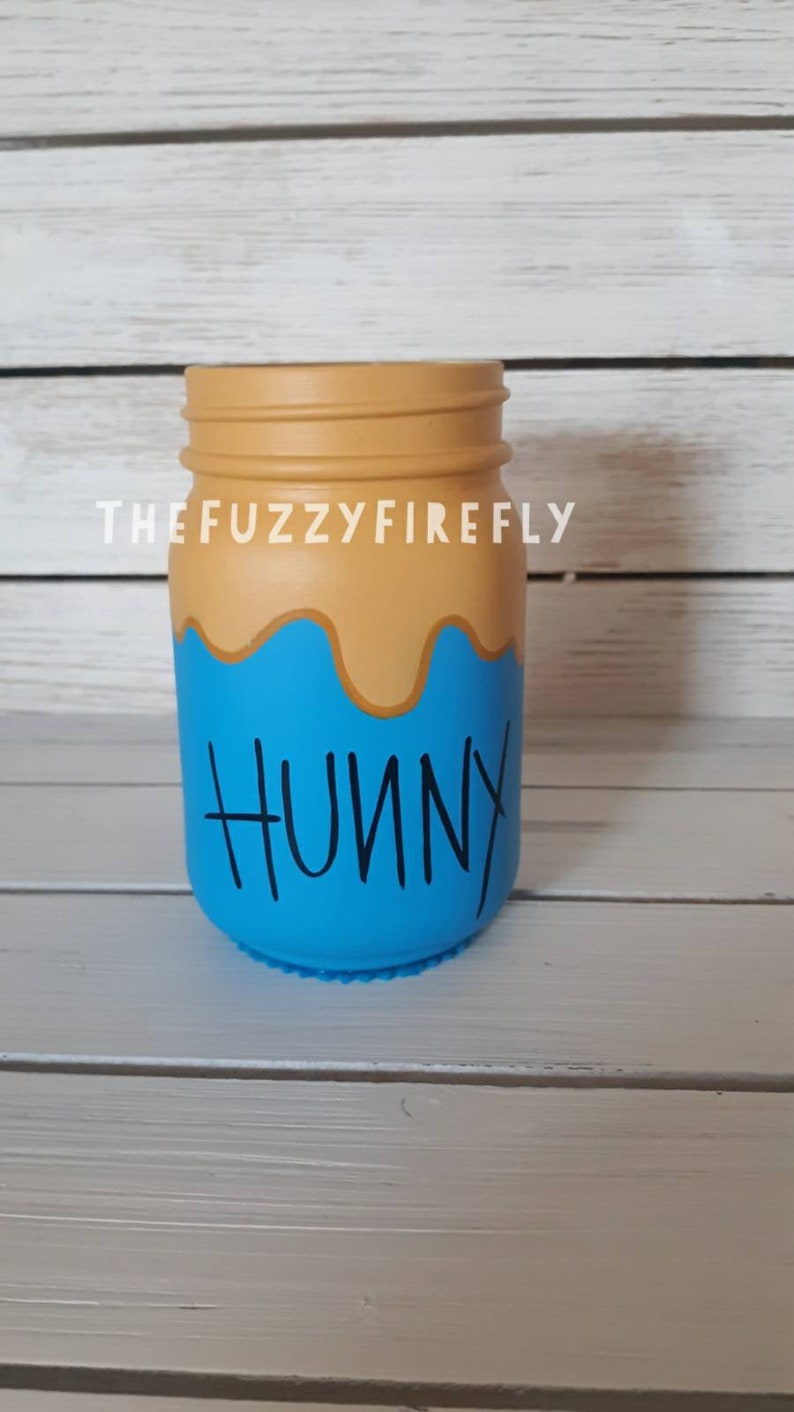 Peut inclure: Un bocal en verre peint avec une section sup&eacute;rieure beige rappelant du miel qui coule et une section inf&eacute;rieure bleu vif. Le mot HUNNY est &eacute;crit en noir dans une police manuscrite sur la partie bleue. Le texte THEFUZZYFIREFLY est visible au-dessus.