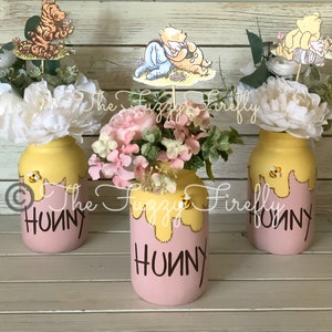 May include: Three glass jars decorated with pink paint and yellow honey dripping down the sides. Each jar has a Winnie the Pooh character topper and is filled with artificial flowers. The jars are labeled "Hunny" in brown lettering.