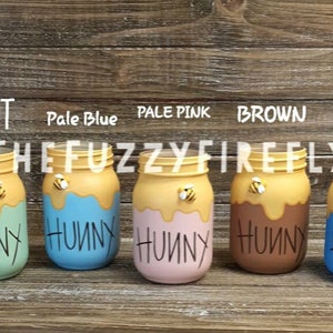 Peut inclure: Cinq pots en verre peints dans diff&eacute;rentes couleurs, notamment menthe, bleu p&acirc;le, rose p&acirc;le, marron et bleu. Chaque pot a un dessus jaune avec un motif de miel qui coule et une petite figurine d'abeille. Les pots sont &eacute;tiquet&eacute;s "Hunny" en lettres noires.