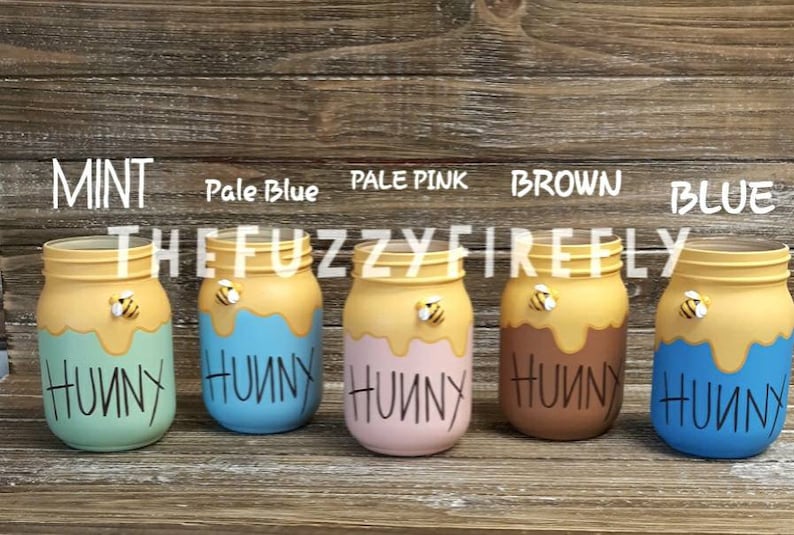 Peut inclure: Cinq pots en verre peints dans diff&eacute;rentes couleurs, notamment menthe, bleu p&acirc;le, rose p&acirc;le, marron et bleu. Chaque pot a un dessus jaune avec un motif de miel qui coule et une petite figurine d'abeille. Les pots sont &eacute;tiquet&eacute;s "Hunny" en lettres noires.